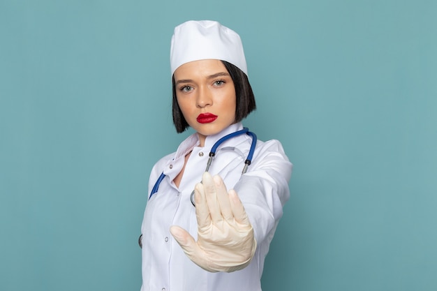 Una vista frontal joven enfermera en traje médico blanco y estetoscopio azul que muestra la señal de stop en el escritorio azul médico médico del hospital