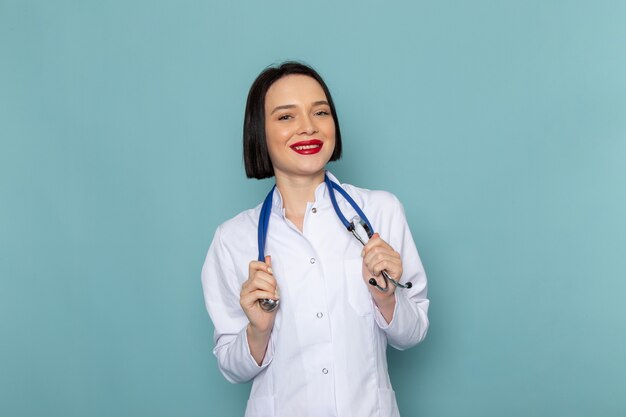 Una vista frontal joven enfermera en traje médico blanco y estetoscopio azul sonriendo en el escritorio azul médico médico del hospital