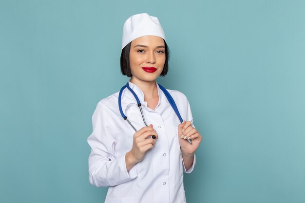 Una vista frontal joven enfermera en traje médico blanco y estetoscopio azul sonriendo en el escritorio azul médico médico del hospital