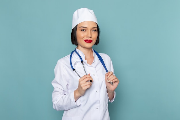 Una vista frontal joven enfermera en traje médico blanco y estetoscopio azul sonriendo en el escritorio azul médico médico del hospital