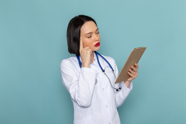 Una vista frontal joven enfermera en traje médico blanco estetoscopio azul sosteniendo el bloc de notas en el escritorio azul médico médico del hospital