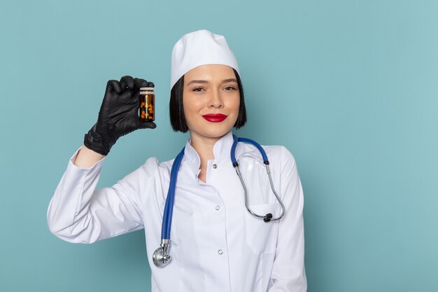 Una vista frontal joven enfermera en traje médico blanco y estetoscopio azul sosteniendo píldoras en el escritorio azul médico médico del hospital