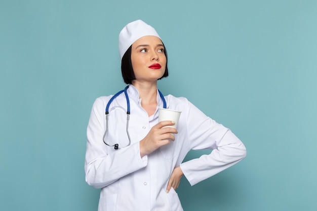 Una vista frontal joven enfermera en traje médico blanco y estetoscopio azul sosteniendo un vaso de plástico en el escritorio azul médico médico del hospital