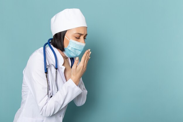 Una vista frontal joven enfermera en traje médico blanco y máscara de estetoscopio azul tosiendo en el escritorio azul médico médico del hospital