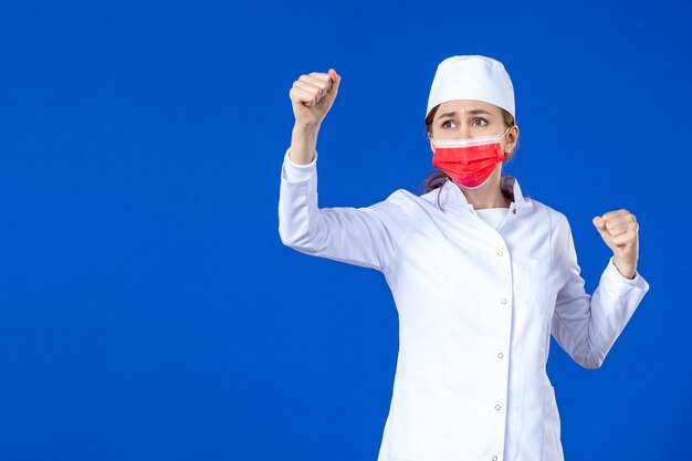 Vista frontal joven enfermera en traje médico con máscara protectora roja en la pared azul