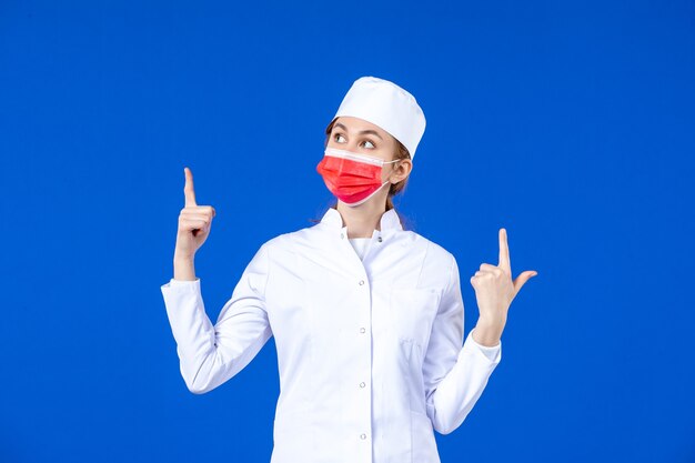 Vista frontal joven enfermera en traje médico con máscara protectora roja en la pared azul