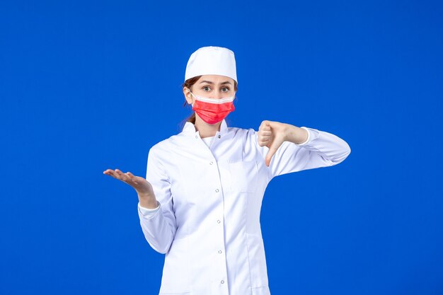 Vista frontal joven enfermera en traje médico con máscara protectora roja en la pared azul