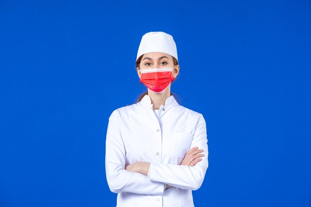 Vista frontal joven enfermera en traje médico con máscara protectora roja en la pared azul