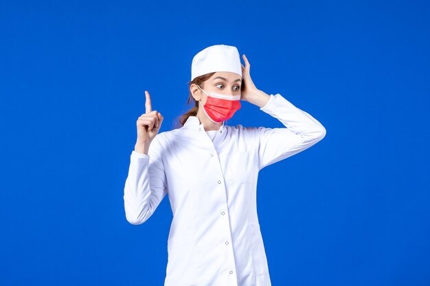 Vista frontal joven enfermera en traje médico con máscara protectora roja en la pared azul