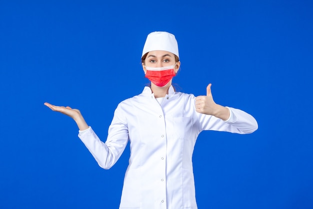 Vista frontal joven enfermera en traje médico con máscara protectora roja en la pared azul