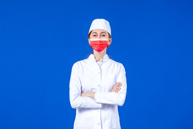 Vista frontal joven enfermera en traje médico con máscara protectora roja en la pared azul