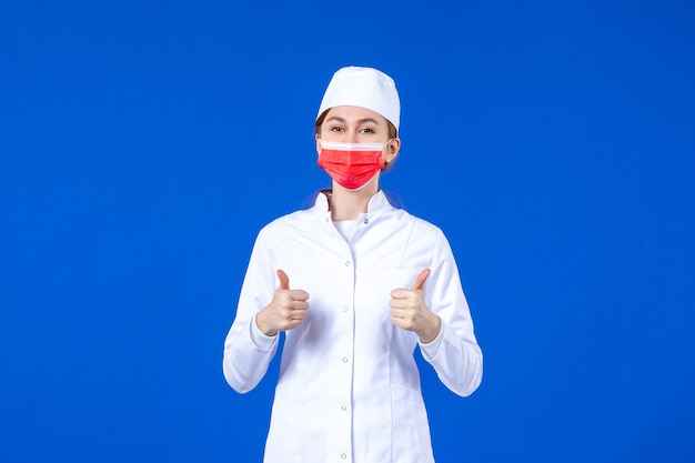 Vista frontal joven enfermera en traje médico con máscara protectora roja en la pared azul