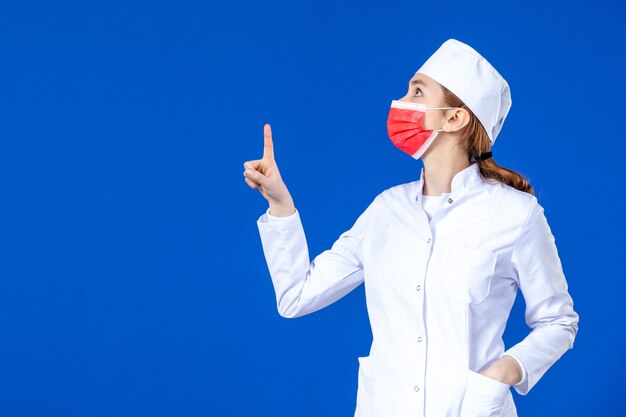 Vista frontal joven enfermera en traje médico con máscara protectora roja en la pared azul