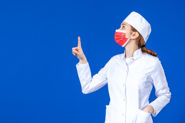 Vista frontal joven enfermera en traje médico con máscara protectora roja en la pared azul