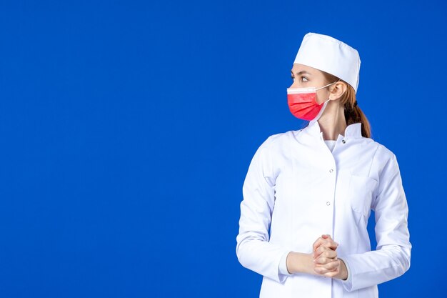 Vista frontal joven enfermera en traje médico con máscara roja sobre pared azul