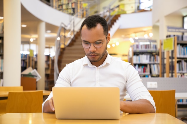 Vista frontal del joven enfocado escribiendo en la computadora portátil en la biblioteca