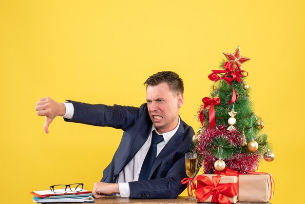 Vista frontal joven enojado haciendo signo de pulgar hacia abajo sentado en la mesa cerca del árbol de Navidad y regalos sobre fondo amarillo
