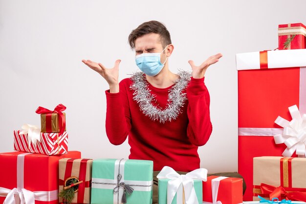 Vista frontal joven enojado con máscara abriendo las manos sentados alrededor de regalos de Navidad