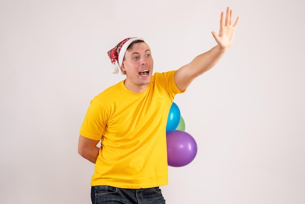 Vista frontal del joven escondiendo globos de colores en la pared blanca