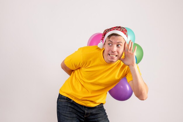 Vista frontal del joven escondiendo globos de colores en la pared blanca