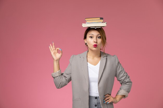 Vista frontal joven estudiante en abrigo gris con libros sobre su cabeza sobre fondo rosa estudio de la universidad de lecciones de fondo