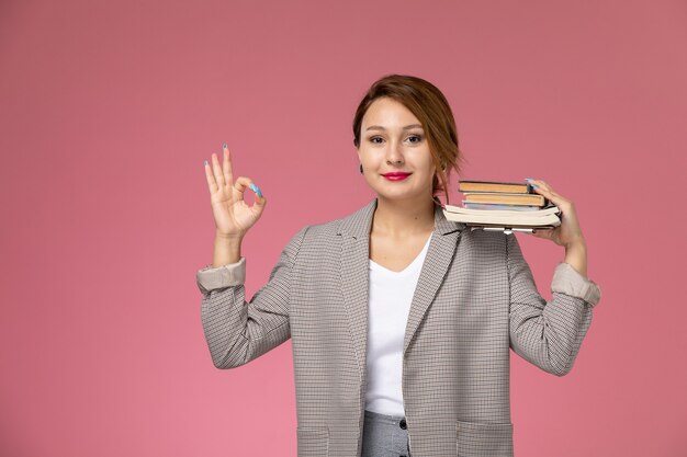 Vista frontal joven estudiante en abrigo gris posando sosteniendo libros y sonriendo en el estudio de la universidad de lecciones de fondo rosa