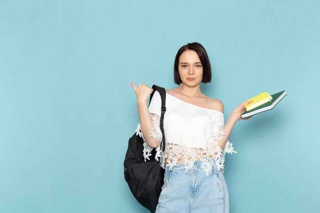 Vista frontal joven estudiante en camisa blanca blue jeans y bolso negro sosteniendo cuadernos en el espacio azul estudiante universitaria enseñar escuela