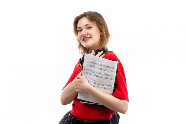 Una vista frontal joven estudiante en camisa roja bolsa negra con auriculares negros sonriendo sosteniendo cuaderno en el blanco