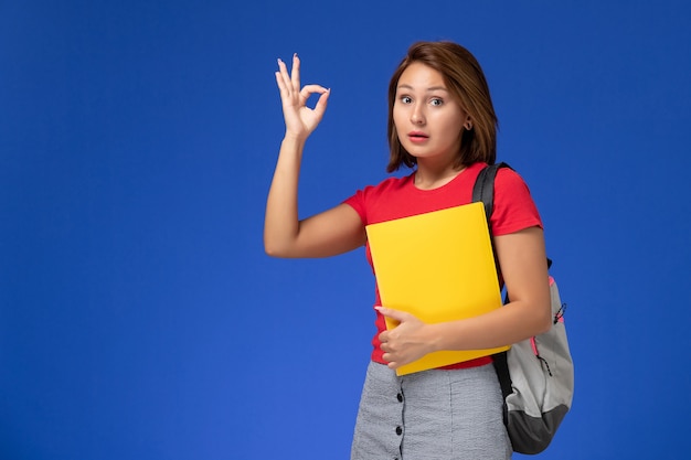 Vista frontal joven estudiante en camisa roja con mochila con archivos amarillos sobre fondo azul claro.