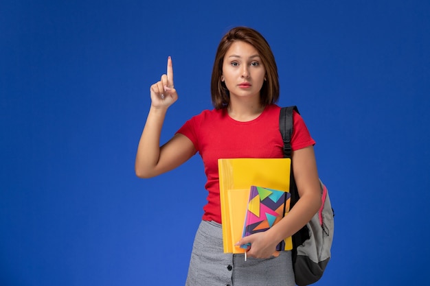 Vista frontal joven estudiante en camisa roja con mochila con archivos y cuaderno sobre el fondo azul claro.