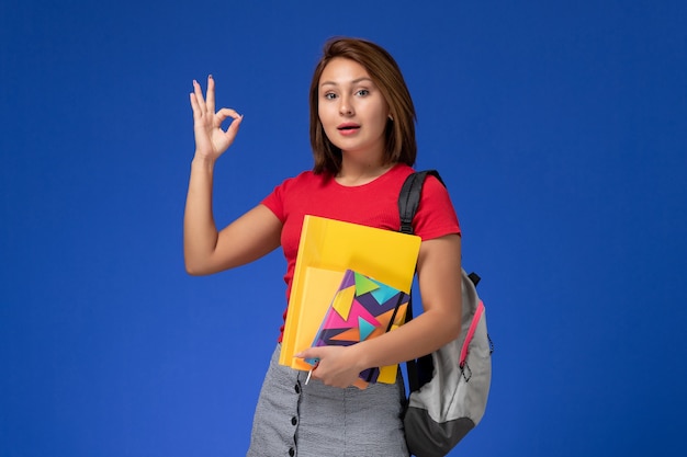 Vista frontal joven estudiante en camisa roja con mochila con archivos y cuaderno sobre fondo azul.