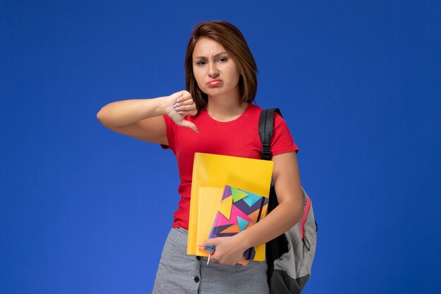 Vista frontal joven estudiante en camisa roja con mochila con archivos y cuaderno sobre fondo azul.