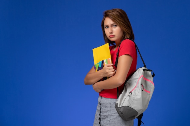 Vista frontal joven estudiante en camisa roja con mochila con cuaderno y archivos sobre fondo azul claro.