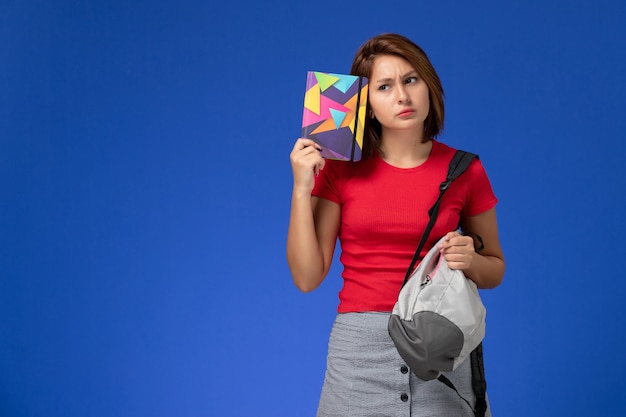 Foto gratuita vista frontal joven estudiante en camisa roja con mochila con cuaderno pensando en fondo azul claro.