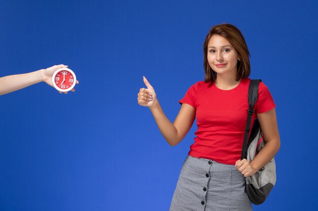 Vista frontal joven estudiante en camisa roja con mochila mostrando como signo sobre fondo azul claro.