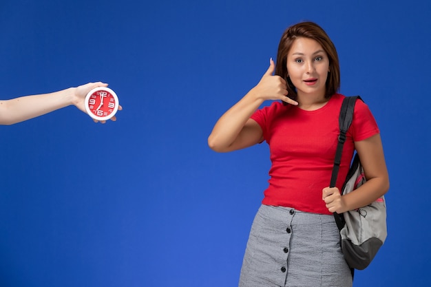 Vista frontal joven estudiante en camisa roja con mochila mostrando pose de llamada telefónica sobre fondo azul claro.