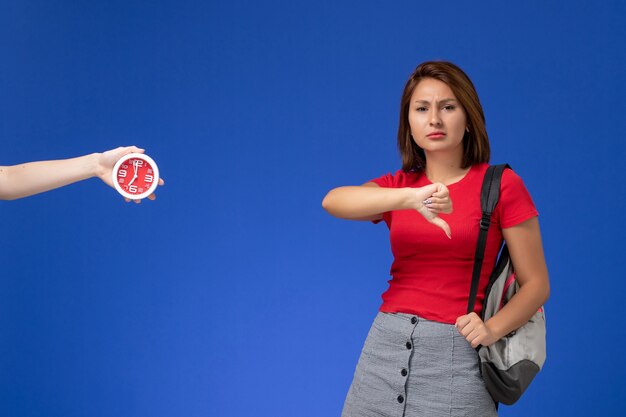 Vista frontal joven estudiante en camisa roja con mochila mostrando signo diferente sobre fondo azul claro.