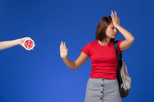 Vista frontal joven estudiante en camisa roja con mochila sobre el fondo azul claro.