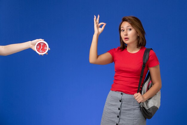 Vista frontal joven estudiante en camisa roja con mochila sobre el fondo azul claro.