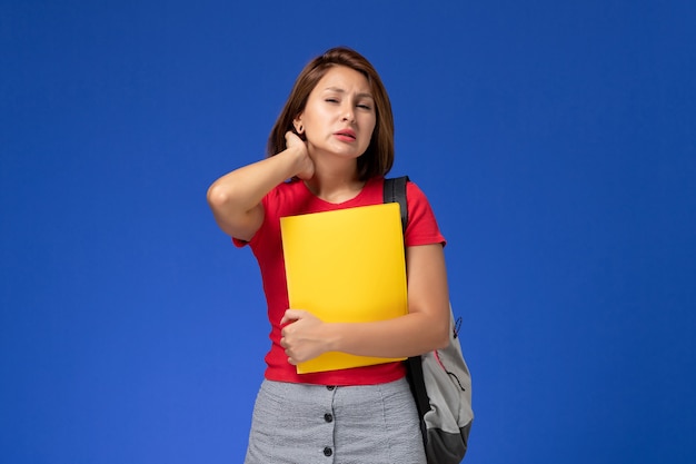 Vista frontal joven estudiante en camisa roja con mochila sosteniendo archivos amarillos con dolor de cuello sobre fondo azul claro.