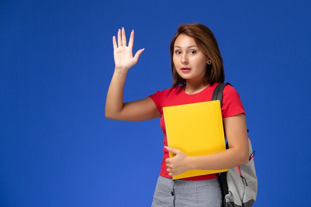 Vista frontal joven estudiante en camisa roja con mochila sosteniendo archivos amarillos ondeando sobre fondo azul claro.