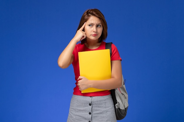 Vista frontal joven estudiante en camisa roja con mochila sosteniendo archivos amarillos y pensando en fondo azul claro.