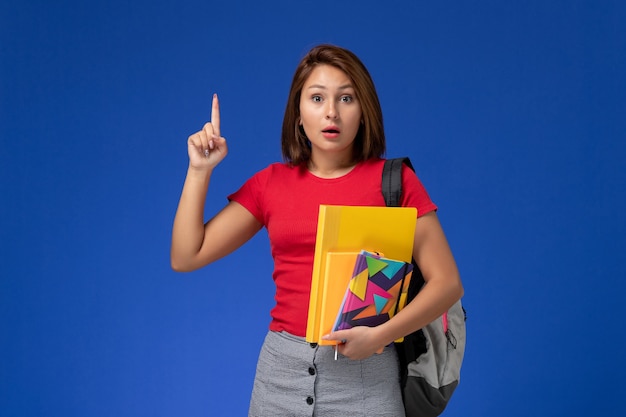 Vista frontal joven estudiante en camisa roja con mochila sosteniendo archivos y cuaderno levantando su dedo sobre el escritorio azul.