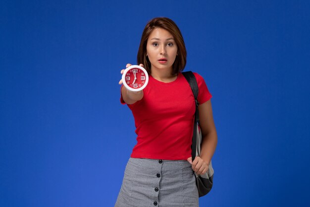 Vista frontal joven estudiante en camisa roja con mochila sosteniendo relojes en el fondo azul claro.