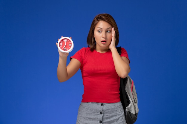 Vista frontal joven estudiante en camisa roja con mochila sosteniendo relojes en el fondo azul claro.