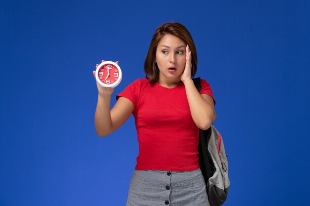 Vista frontal joven estudiante en camisa roja con mochila sosteniendo relojes en el fondo azul claro.