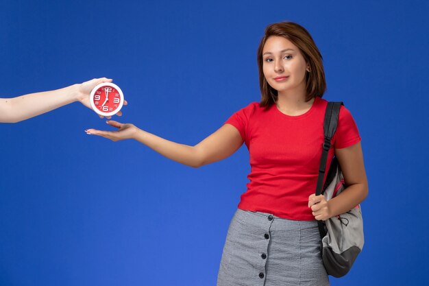 Vista frontal joven estudiante en camisa roja con mochila sosteniendo relojes sonriendo sobre fondo azul claro.