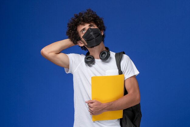 Vista frontal joven estudiante en camiseta blanca con máscara negra y sosteniendo archivos amarillos pensando en fondo azul claro.