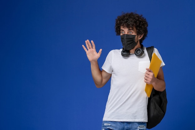 Foto gratuita vista frontal joven estudiante en camiseta blanca con máscara negra y sosteniendo archivos amarillos sobre el fondo azul claro.