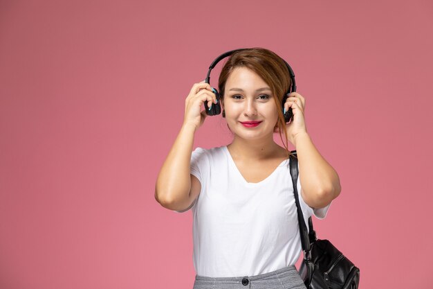 Vista frontal joven estudiante en camiseta blanca y pantalón gris con auriculares sobre fondo rosa lecciones estudiantiles universidad universitaria